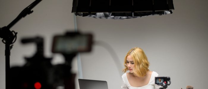Behind the scenes young blonde woman entrepreneur working with laptop presents cosmetic products during online live stream over white background studio, selling online and beauty blogger concept
